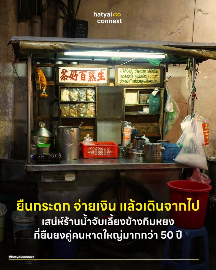 ยืนกระดก จ่ายเงิน แล้วเดินจากไป เสน่ห์ร้านน้ำจับเลี้ยงข้างกิมหยงที่ยืนยงคู่คนหาดใหญ่มากกว่า 50 ปี . เมื่อแสงไฟจากสี่แยกโรงแรมวี.แอล. สาดส่องลงบนถนนเพชรเกษมยามค่ำคืน ภาพผู้คนหลากหลายวัยจอดรถแวะเวียนมายืนมุงรถเข็นคันเล็กๆ หน้าธนาคารกสิกรไทยสาขาตลาดกิมหยงคือฉากทัศน์ที่อยู่คู่เมืองหาดใหญ่มานานกว่าครึ่งศตวรรษ . สำหรับผู้ใหญ่วัยเก๋านี่คือโอเอซิสแห่งความสดชื่นที่คุ้นเคย แต่สำหรับคนรุ่นใหม่จำนวนมาก น้ำสีดำเข้มในแก้วใสอาจเป็นปริศนาที่ไม่เคยทำความรู้จัก เบื้องหลังกลับซ่อนมิติทางสังคม วัฒนธรรมและการเชื่อมโยงผู้คนในเมืองเศรษฐกิจแห่งนี้ไว้อย่างลึกซึ้ง . #ปริศนาโอสถสีดำและสารพัดชื่อเรียก ความคลาสสิกแรกของร้านนี้คือการเป็นตำนานที่ไม่มีป้ายชื่อร้านตายตัว ผู้คนต่างเรียกขานกันตามความทรงจำ บ้างเรียกว่า 'ร้านน้ำสมุนไพรข้างตลาดกิมหยง' บ้างเรียก 'ร้านน้ำจับเลี้ยงกสิกร' หรือบางคนก็จำในชื่อคนขายอย่าง 'ร้านโกอ๊อด' แต่สิ่งที่ทุกคนรู้ใจกันคือเมนูน้ำสมุนไพรสูตรโบราณ ที่แบ่งออกเป็น 2 ขั้วรสชาติ . แก้วแรกคือ 'น้ำเลี้ยงจุ้ย' หรือที่คนทั่วไปเรียกว่าน้ำจับเลี้ยง หอมหวาน ดื่มง่าย ช่วยดับกระหายและแก้ร้อนใน ไฮไลต์ที่แท้จริงคือ 'น้ำขม' น้ำสีดำที่รสชาติที่เด็กยุคใหม่อาจเบือนหน้าหนี รวมๆ แล้วเครื่องดื่มประเภทนี้มีหลายรสชาติ หลายเลเวลความขม และมีหลายชื่อเรียกมาก แต่ขอเรียกรวมว่า 'น้ำขม' เพราะเป็นตัวบอสขั้นสุดยอดของสรรพคุณและรสชาติ ที่สำคัญคือเป็นชื่อที่รู้จักในวงกว้าง ในภาษาจีนกลางเรียกว่า 'ขู่ฉา' แปลตรงตัวก็คือน้ำขมนั่นเอง . ซึ่งมีส่วนผสมของสมุนไพรมากมาย ถือเป็นคัมภีร์ลับสูตรใครสูตรมัน ในความขมนั้นอัดแน่นไปด้วยสรรพคุณการถอนพิษร้อนในร่างกาย การลองดื่มน้ำขมสักแก้วจึงเปรียบเสมือนบททดสอบความกล้าและก้าวแรกของการเข้าสู่วิถีคนหาดใหญ่ดั้งเดิม . #สถาปัตยกรรมสตรีตฟูดและพื้นที่แห่งความเท่าเทียม หากมองในมุมมองทางสังคมวิทยา รูปแบบการขายของร้านนี้คือตัวแทนของวัฒนธรรมสตรีตฟูดแบบ Stall (แผงลอยแบบไม่มีที่นั่ง) อย่างแท้จริง การไม่มีโต๊ะเก้าอี้ให้นั่งจับกลุ่มสังสรรค์ได้สร้างพฤติกรรมการบริโภคที่มีเอกลักษณ์คือ 'ยืนกระดก จ่ายเงิน แล้วเดินจากไป' ลูกค้าเพียงแค่เดินเข้ามา หยิบแก้วที่รินเตรียมไว้ ดื่มรวดเดียว วางแก้ว จ่ายเงิน แล้วเดินทางต่อ . วิถีการบริโภคที่รวดเร็วและเป็นกันเองเช่นนี้ ชวนให้จินตนาการถึงวัฒนธรรมการดื่มกาแฟเอสเปรสโซ (Espresso) แบบยืนดื่มหน้าบาร์ในอิตาลี ที่ผู้คนแวะมาเติมพลังช็อตเข้มๆ ดื่มรวดเดียวจบแล้วไปต่อ เพียงแต่เปลี่ยนจากคาเฟอีนรสเข้มในยุโรป มาเป็นน้ำสมุนไพรใจกลางเมืองหาดใหญ่ . พื้นที่หน้าแผงรถเข็นเล็กๆ นี้จึงทำหน้าที่เป็นพื้นที่ทางสังคมที่ไร้ชนชั้น ในช่วงเวลาไม่กี่วินาทีที่ยืนดื่ม ทุกคนถูกดึงให้มาอยู่บนระนาบเดียวกันผ่านแก้วน้ำสมุนไพรราคาไม่แพง ถือเป็นการทลายกำแพงทางสังคมและเป็นสะพานเชื่อมช่องว่างระหว่างเจเนอเรชันที่คาเฟ่ยุคปัจจุบันอาจให้ไม่ได้ . #ปฏิบัติการอาสาสมัครสายแดกปลุกชีวิตเมือง เพื่อเป็นการต่อลมหายใจให้ตำนานและปลุกเมืองหาดใหญ่ให้กลับมามีชีวิตชีวา ทางเพจ แอบแดก ได้ผุดแคมเปญกระตุ้นเศรษฐกิจสุดสร้างสรรค์ โดยตั้งขบวน 'อาสาสมัครสายแดก' นัดรวมพลคนรักเมืองหาดใหญ่มาช่วยกันอุดหนุนร้านท้องถิ่น ประเดิมคิวแรกกันที่ร้านน้ำจับเลี้ยงหน้ากสิกรแห่งนี้ . ความน่าสนใจคือกิจกรรมนี้ดึงเอากลยุทธ์แบบคนรุ่นใหม่มาผสมผสานกับความคลาสสิกของร้านได้อย่างลงตัว ด้วยการสร้างชาเลนจ์ให้ผู้เข้าร่วมถ่ายรูปรีแอกชันตอนกระดกน้ำขมแบบเรียลๆ ฮาๆ (ใครไม่ไหวไม่บังคับ) พร้อมแจกเข็มกลัดพิเศษทีมถล่มแดกรุ่นก่อตั้งให้กับลูกเพจที่ไปแสดงตัวหน้าร้าน . ตัวอย่างที่ทรงพลังของการใช้สื่อสังคมออนไลน์เพื่อสร้างแรงกระเพื่อมในโลกแห่งความเป็นจริง ช่วยกระจายรายได้สู่ผู้ประกอบการรายย่อย และยังช่วยแนะนำวัฒนธรรมดั้งเดิมให้กลับมาป๊อปในหมู่คนรุ่นใหม่อีกครั้ง หากใครกิจกรรมรวมพลแบบนี้ สามารถติดตามเพจแอบแดกเพื่อดูการนัดครั้งต่อไปได้เลย . #มนต์ขลังแห่งความเรียบง่ายที่ไร้การปรุงแต่ง ในยุคที่คาเฟ่และร้านอาหารต่างแข่งขันกันตกแต่งสถานที่ให้สวยงามเพื่อดึงดูดสายตา ร้านรถเข็นลักษณะนี้กลับเปี่ยมไปด้วยความขลังที่เกิดจากการหยุดกาลเวลาเอาไว้ รถเข็นคันเก่า แสงไฟสลัว แก้วใสที่วางซ้อนกันเรียงราย รอรับการรินน้ำสีดำ ทั้งหมดนี้คือความดิบและความแท้ที่ไม่ได้เกิดจากการจัดฉากหรือทำการตลาด เกิดจากการยืนหยัดขายสิ่งเดิม ณ จุดเดิมมานานกว่าครึ่งศตวรรษ ความขลังจึงไม่ได้อยู่ที่ความหรูหรา อยู่ที่ความสม่ำเสมอ กลิ่นสมุนไพรที่ต้มจนได้ที่และเสียงแก้วกระทบกันที่ยังคงดังกังวานเหมือนเมื่อ 50 ปีก่อน . พิกัดร้าน สถานที่ : ถนนเพชรเกษม หน้าธนาคารกสิกรไทย (ข้างตลาดกิมหยง) ตรงข้ามร้านเซาะฮึ้ง เวลาเปิด-ปิด : 18:30 - 23:00 น. __ #HatyaiConnext #น้ำจับเลี้ยงหาดใหญ่ #โกอ๊อดกิมหยง #รีวิวหาดใหญ่ #มิติสังคมเมือง #หาดใหญ่hatyaiconnext